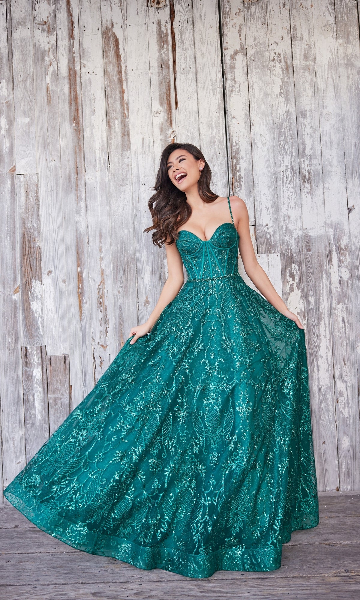 A woman poses in front of a weathered wooden wall, holding out the skirt of her Colette Glitter-Tulle Long Prom Ball Gown CL5117.